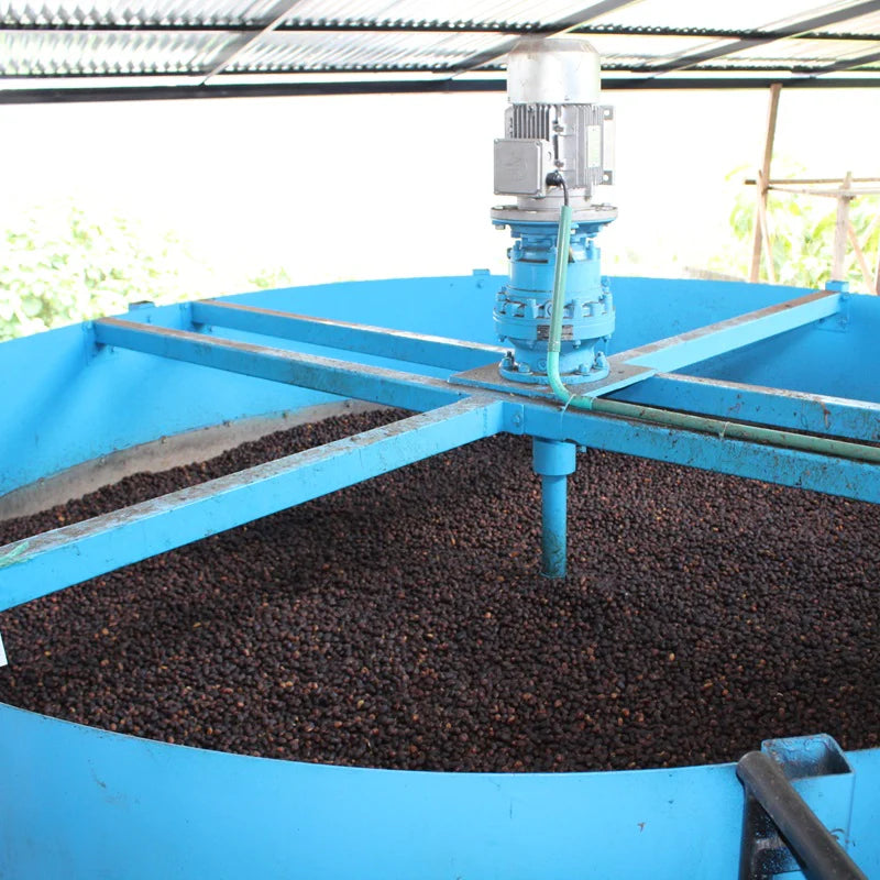 Blue cylindrical machine with a central shaft processing dark material, likely coffee beans, outdoors.