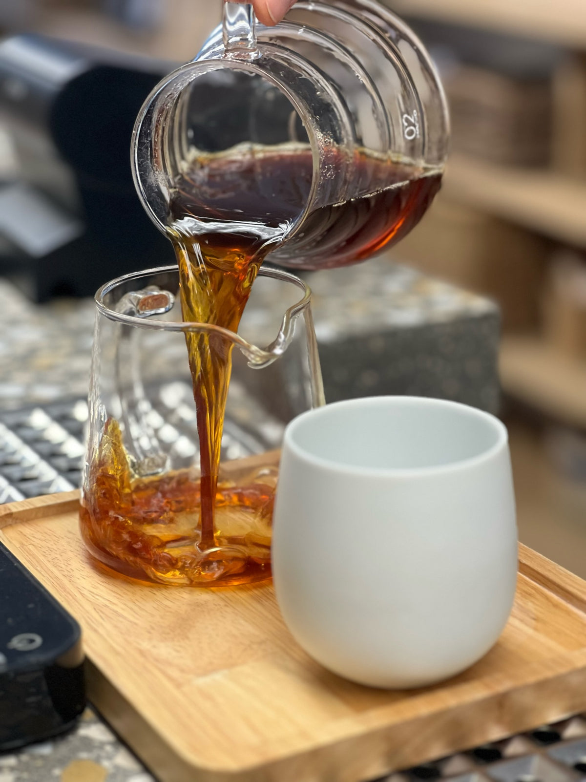Hario Glass Server pouring coffee into a glass server and a white mug on a wooden tray.